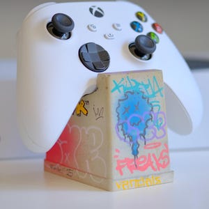 May include: White Xbox controller on a graffiti-style stand. The stand is beige with pink, blue, and yellow spray-painted designs and the words "FREAKS" and "VANDALS".