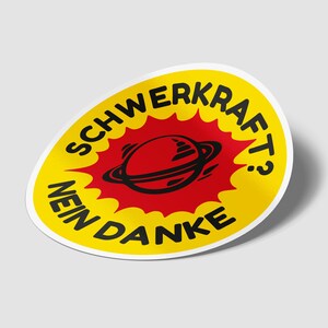 Puede incluir: Una pegatina redonda amarilla y roja con el texto "SCHWERKRAFT? NEIN DANKE" y un planeta de dibujos animados negro y rojo con un anillo a su alrededor.