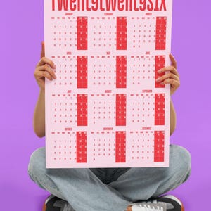 May include: A pink 2026 calendar with red text and calendar boxes. The calendar is held by a person sitting cross-legged, wearing blue jeans and trainers, against a purple background. The calendar's title is "TWENTYTWENTYSIX".