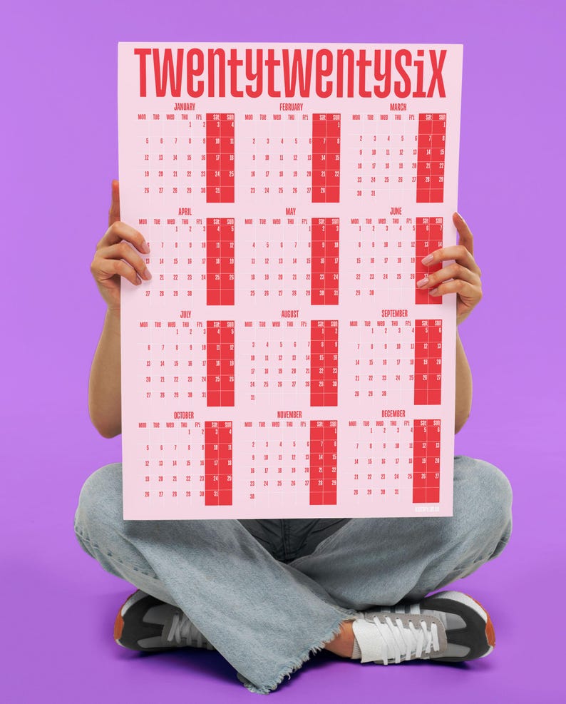 May include: A pink 2026 calendar with red text and calendar boxes. The calendar is held by a person sitting cross-legged, wearing blue jeans and trainers, against a purple background. The calendar's title is "TWENTYTWENTYSIX".