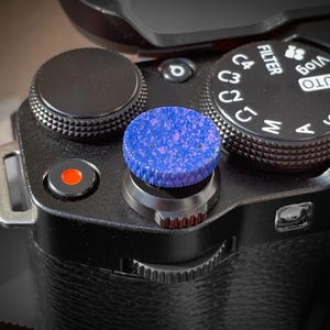 May include: Close-up of a black camera with a blue and purple textured shutter button. The camera has various dials and buttons, including a mode dial with settings like "AUTO" and "Vlog". The camera body is black with a textured grip.