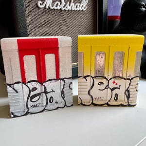 Puede incluir: Dos pequeñas cajas rectangulares pintadas a mano. Una es blanca con un diseño de puerta roja, la otra es amarilla con un diseño de ventana. Ambas presentan letras de estilo grafiti en blanco y negro en la parte inferior. Las cajas están sobre una superficie blanca.
