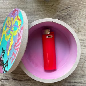 May include: Open concrete container with a lid decorated with colourful graffiti art. A red BIC lighter is inside, against a pink interior. The round container sits on a wooden surface.