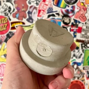 Puede incluir: Una tapa de bote de pintura en aerosol de hormigón gris claro con una boquilla circular y una pequeña letra grabada. La tapa se sostiene en una mano sobre un fondo de pegatinas coloridas.