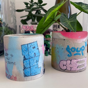 Może przedstawiać: Dwie cylindryczne betonowe doniczki z kolorowymi grafikami w stylu graffiti. Na lewej doniczce widnieje czarny napis "ART IS NOT A CRIME". Na prawej doniczce znajduje się niebieski napis "soup" oraz różowe litery tworzące słowo "CHE".