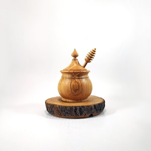 May include: A wooden honey pot with a lid and a wooden honey dipper. The honey pot is sitting on a wooden base.