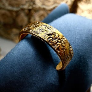 Puede incluir: Un brazalete dorado con un intrincado diseño floral en relieve.