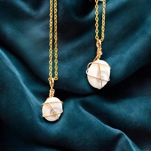 Peut inclure: Deux colliers dorés avec pendentifs. Chaque pendentif présente une pierre blanche de forme ovale enveloppée dans du fil d'or. Les colliers sont posés sur un fond en tissu bleu canard foncé.