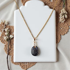 Puede incluir: Un collar de cadena dorada con un colgante ovalado gris oscuro. El colgante tiene un patrón moteado y está suspendido de una fianza dorada. El collar se exhibe en un expositor de joyas blanco.