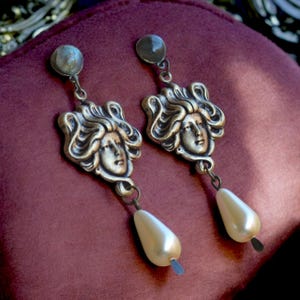 May include: A pair of silver tone earrings with a pearlescent drop and a detailed face design.
