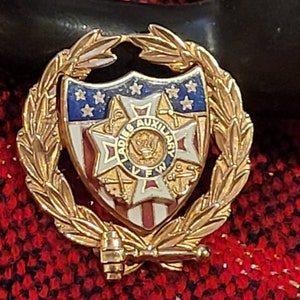 May include: Gold-toned pin with a wreath of leaves surrounding a shield with an American flag design. The shield also features a red, white, and blue cross with the words "Ladies Auxiliary VFW of the US" in a circle around it.