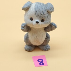 May include: A ceramic figurine of a puppy with a white face and gray ears, paws, and body. The puppy has black eyes and is holding its paws up. A pink sticky note with the number 8 is at the bottom.