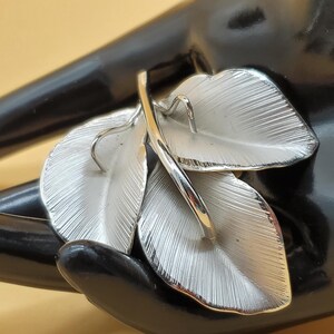 Puede incluir: Broche de metal plateado con tres hojas estilizadas. Las hojas son texturizadas y tienen un diseño ligeramente en relieve.