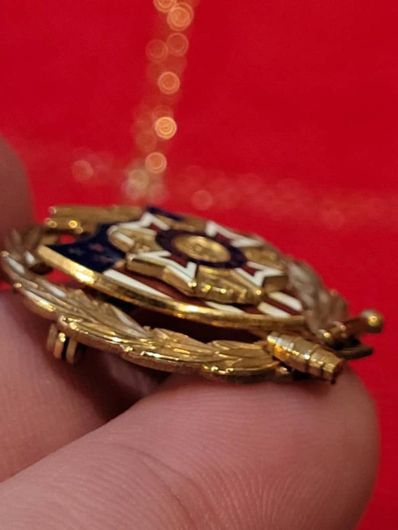 Puede incluir: Un pin de tono dorado con un dise&ntilde;o de escudo rojo, blanco y azul. El escudo tiene una estrella en el centro y est&aacute; rodeado por una corona de hojas doradas. El pin tiene un cierre de pin.