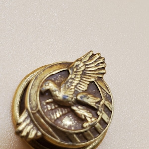 May include: A gold-toned metal button with a raised design of a bird in flight. The bird is surrounded by a circle with a striped pattern.