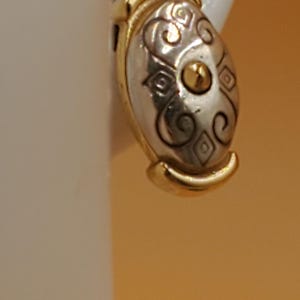 May include: Oval-shaped earring with a silver-tone center and gold-tone trim. The silver section features an ornate design with swirls and diamond shapes, and a gold accent in the center. The earring is set against a neutral background.