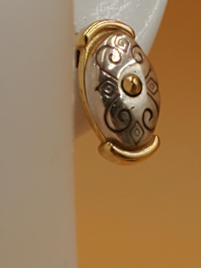 May include: Oval-shaped earring with a silver-tone center and gold-tone trim. The silver section features an ornate design with swirls and diamond shapes, and a gold accent in the center. The earring is set against a neutral background.