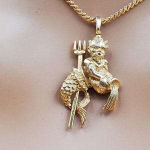 May include: Gold-toned pendant necklace featuring a mythical creature with a trident. The pendant depicts a seated figure with a fish tail, holding a trident. The necklace has a rope chain. The pendant and chain are gold in color.