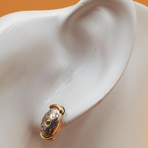 May include: Oval-shaped stud earring with a gold-tone frame and a silver-tone center. The center features a decorative pattern with a small gold-tone circle in the middle. The earring is displayed on a white surface.
