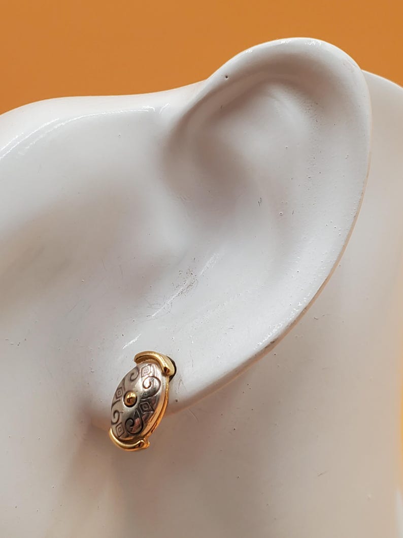 May include: Oval-shaped stud earring with a gold-tone frame and a silver-tone center. The center features a decorative pattern with a small gold-tone circle in the middle. The earring is displayed on a white surface.