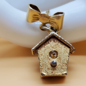 May include: Gold-toned earring featuring a small birdhouse charm with a round, textured detail in the center. The birdhouse is attached to a gold-toned bow.
