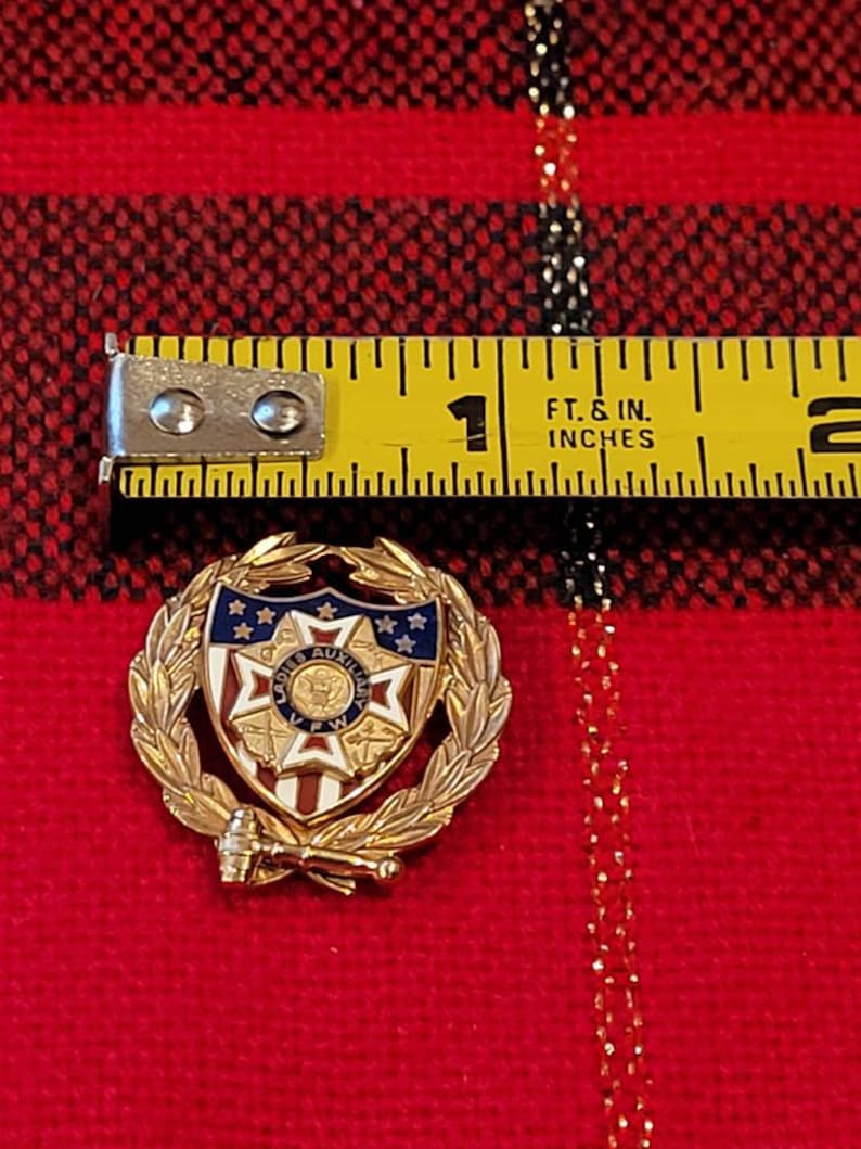 Puede incluir: Pin de color dorado con un escudo rojo, blanco y azul con una estrella en el centro. El escudo est&aacute; rodeado por una corona de hojas doradas. El texto "Ladies Auxiliary VFW" est&aacute; en el escudo. El pin tiene aproximadamente 1 pulgada de di&aacute;metro.