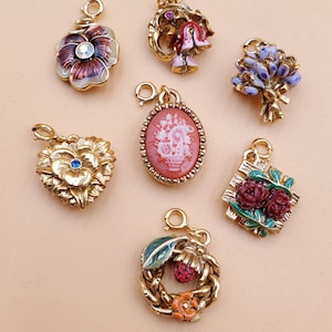 May include: A selection of gold-coloured metal charms with diverse designs, including flowers, a cameo, and a wreath. The charms feature colourful enamel details in shades of red, purple, blue, and green, and are adorned with small gemstones. Each charm has a loop for attaching to a necklace or bracelet.