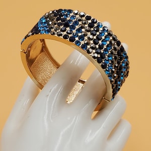 May include: A gold bangle bracelet with a wide band of blue and black rhinestones. The bracelet is open and has a hinged clasp.