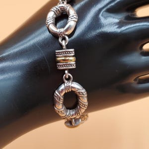 Op de afbeelding: Zilveren armband met twee ovale schakels met textuur en een goudkleurig rechthoekig middendeel. De armband wordt getoond op een zwart oppervlak.