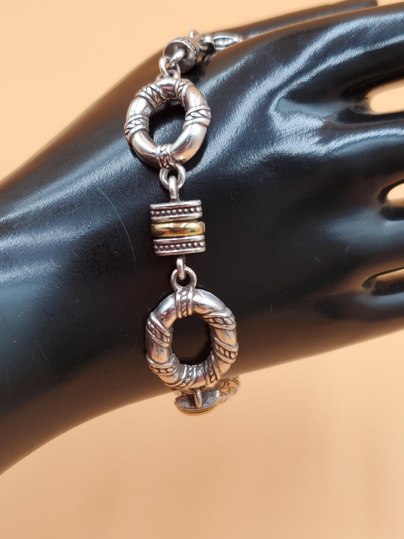 Op de afbeelding: Zilveren armband met twee ovale schakels met textuur en een goudkleurig rechthoekig middendeel. De armband wordt getoond op een zwart oppervlak.