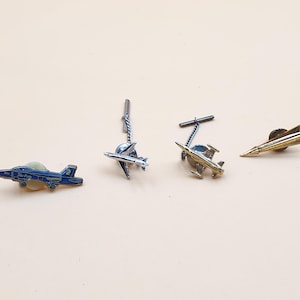 Puede incluir: Colección de pines y pasadores de corbata con temática de aviación. Incluye un pin de avión azul y blanco, dos pasadores de corbata de avión y un pin de cohete dorado. Los artículos se muestran sobre una superficie beige claro.