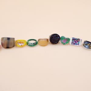 May include: A collection of colorful rings in various designs. The rings include a striped ring, a brown ring, a yellow ring with a bird, a green ring with the word "love", a clear ring, a black rose ring, a green heart ring, a mosaic ring, and two black rings.