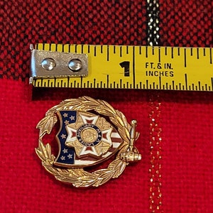 Puede incluir: Pin de tono dorado con un dise&ntilde;o de escudo que presenta una bandera estadounidense roja, blanca y azul, una estrella y las palabras "Auxiliary Ladies of M.V.A.". El escudo est&aacute; rodeado por una corona de hojas doradas.