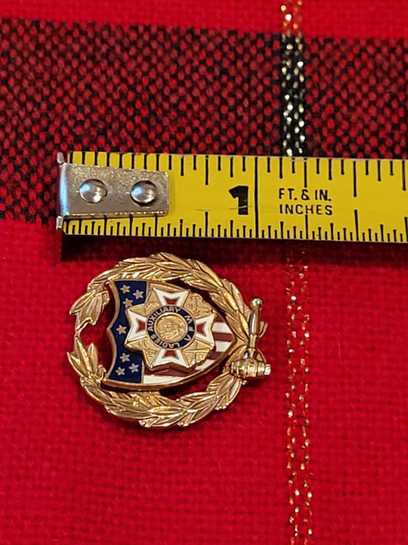 Puede incluir: Pin de tono dorado con un dise&ntilde;o de escudo que presenta una bandera estadounidense roja, blanca y azul, una estrella y las palabras "Auxiliary Ladies of M.V.A.". El escudo est&aacute; rodeado por una corona de hojas doradas.