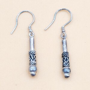 May include: A pair of silver dangle earrings. Each earring features a long, cylindrical body with a decorative, patterned band and a small, rounded bead at the bottom. The earrings have a simple hook design for pierced ears.