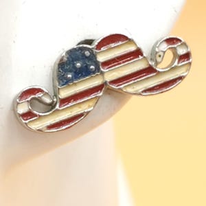 May include: A patriotic mustache-shaped earring with a silver-colored outline. The earring features the American flag design, with red and white stripes and a blue field with white stars. The earring is a unique accessory.