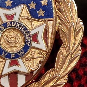 Puede incluir: Pin de tono dorado con un escudo rojo, blanco y azul con una estrella y un ancla. El escudo est&aacute; rodeado por una corona de hojas doradas. El texto "US AUXILIARY VFW" es visible en el escudo.