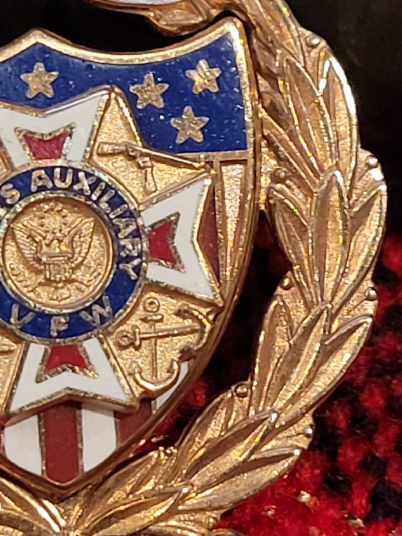 Puede incluir: Pin de tono dorado con un escudo rojo, blanco y azul con una estrella y un ancla. El escudo est&aacute; rodeado por una corona de hojas doradas. El texto "US AUXILIARY VFW" es visible en el escudo.