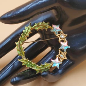 May include: A gold-toned brooch featuring a wreath design with green enamel leaves and iridescent star accents. The brooch has a pin closure on the back. A decorative jewelry piece.