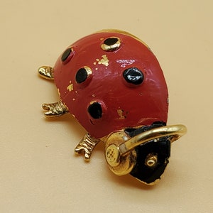 May include: A ladybug brooch with a red body, black spots, and gold accents. The brooch has a gold-colored head and legs. The ladybug's body has a distressed, vintage appearance. The brooch is set against a light beige background.