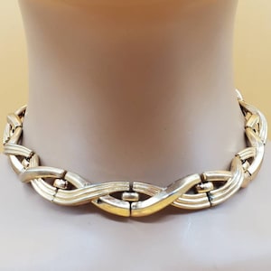 May include: A gold-toned metal choker necklace with a woven design. The necklace is made of interlocking links that create a wavy pattern.