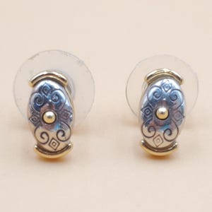 May include: A pair of silver-tone stud earrings with gold-tone accents. Each earring features an oval design with intricate patterns and a small gold-tone center. The earrings have clear plastic backings.