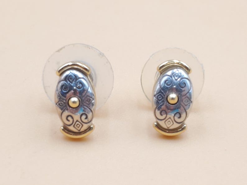 May include: A pair of silver-tone stud earrings with gold-tone accents. Each earring features an oval design with intricate patterns and a small gold-tone center. The earrings have clear plastic backings.