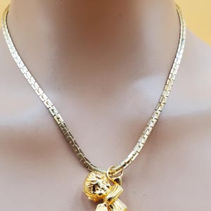 Pu&ograve; includere: Una collana dorata con un ciondolo a forma di cherubino che tiene un arco e una freccia. Il ciondolo &egrave; sospeso a una catena a maglie piatte e rettangolari. La collana &egrave; esposta su un manichino.