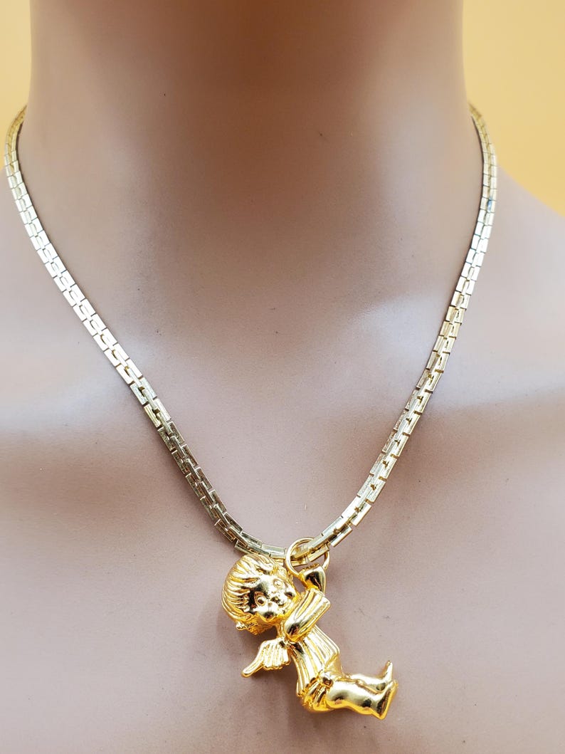 Pu&ograve; includere: Una collana dorata con un ciondolo a forma di cherubino che tiene un arco e una freccia. Il ciondolo &egrave; sospeso a una catena a maglie piatte e rettangolari. La collana &egrave; esposta su un manichino.