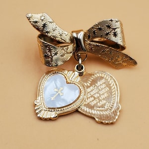Puede incluir: Broche dorado con diseño de lazo y detalles florales. Debajo del lazo cuelga un colgante en forma de corazón con incrustaciones de nácar y una cruz. El colgante está unido a una segunda pieza en forma de corazón con texto grabado.