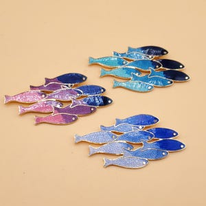 May include: Three clusters of fish-shaped enamel pins with gold-toned outlines. One cluster features a pink to purple gradient, another shows a light blue to dark blue transition, and the third displays a mix of light and dark blue shades.