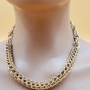 May include: A gold-toned chain necklace with a woven design. The necklace is displayed on a mannequin, showcasing its intricate links and overall style. The chain is a bright, shiny gold color.