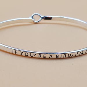 May include: A silver-colored bangle bracelet with the engraved text "IF YOU'RE A BIRD, I'M A BIRD". The bracelet has a heart-shaped clasp. The jewelry is displayed on a light brown surface.