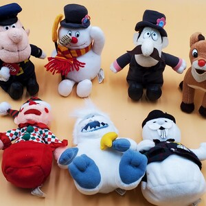 May include: A collection of plush Christmas characters, including a snowman with a scarf, a reindeer, and a police officer. The toys are made of soft materials and feature detailed designs and vibrant colors.